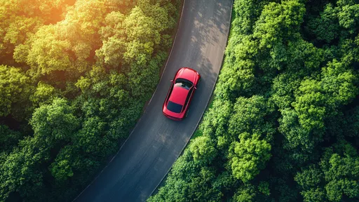 Sarkana automašīna brauc pa meža ceļu, saules gaismā apspīdēta koku ainava.