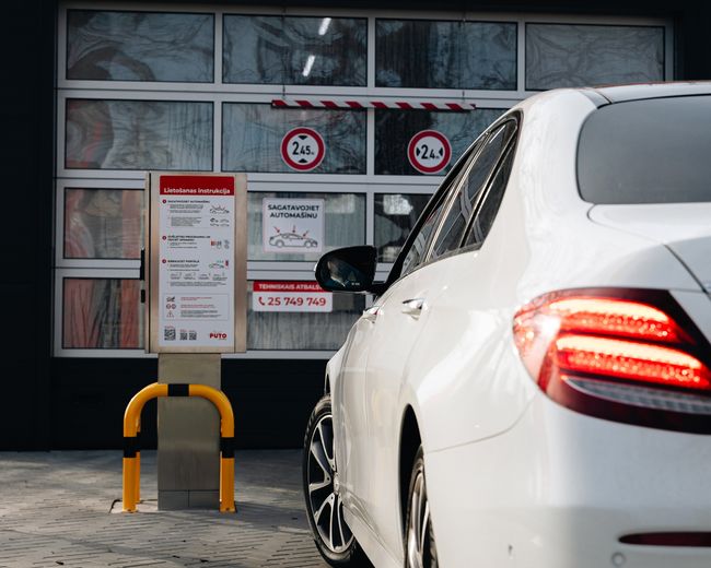 Balta automašīna piebrauc pie automazgātavas ieejas ar zīmēm.