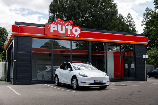 Balta Tesla Model Y pie PUTO Express automātiskās automazgātavas ēkas ar sarkano izkārtni