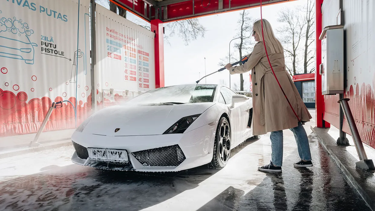 Sieviete pašapkalpošanās automazgātavā ar augstspiediena pistoli mazgā baltu sporta auto.