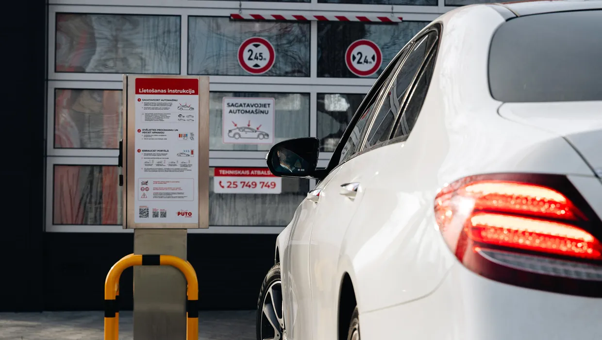 Balta automašīna piebrauc pie automazgātavas ieejas ar zīmēm.